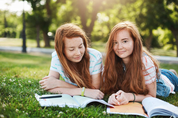 Friendship, family and lifestyle concept. Two attractive redhead girls relaxing on sunny day, lying on grass and doing homework together, smiling at camera with carefree expression - Photo, Image