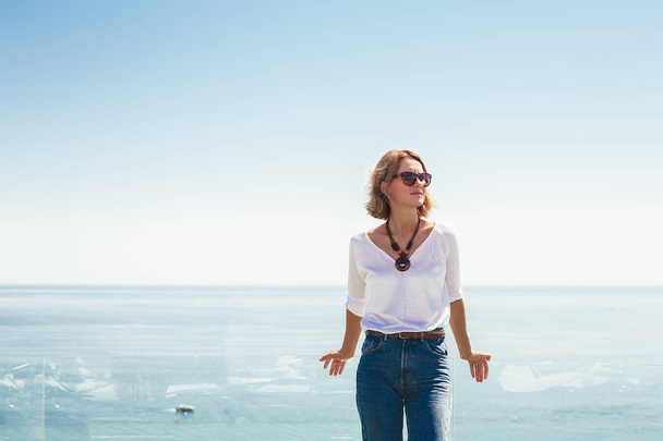 ulkona muotokuva tyylikäs blondi nainen yllään aurinkolasit, valkoinen pusero ja farkut seisoo sininen meri ja taivas tausta
 - Valokuva, kuva