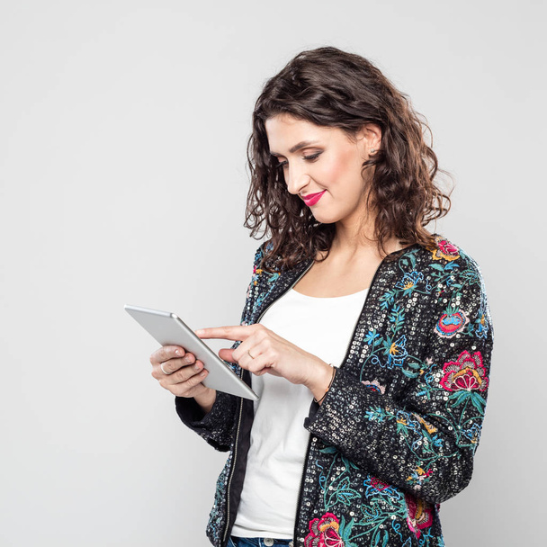 Portrait of beautiful young woman wearing bomber jacket, using a digital tablet. Studio shot, grey background. - Фото, изображение