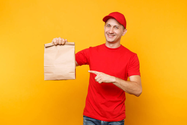 Homme en bonnet rouge, t-shirt donnant ordre de restauration rapide isolé sur fond jaune. Un messager d'employé masculin tient un paquet de papier vide avec de la nourriture. Livraison de produits du magasin ou du restaurant à la maison. Espace de copie - Photo, image