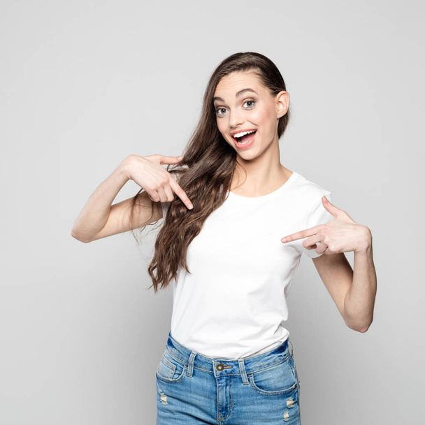 Portrait of excited and happy teenager girl pointing with index fingers at white t-shirt. Studio shot against grey background. - Fotoğraf, Görsel