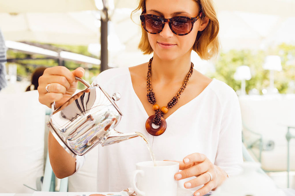 κομψό ξανθιά γυναίκα ρίχνει τσάι στο φλιτζάνι τσαγιέρα εκμετάλλευση κάθεται σε τραπέζι σε υπαίθρια καφετέρια - Φωτογραφία, εικόνα