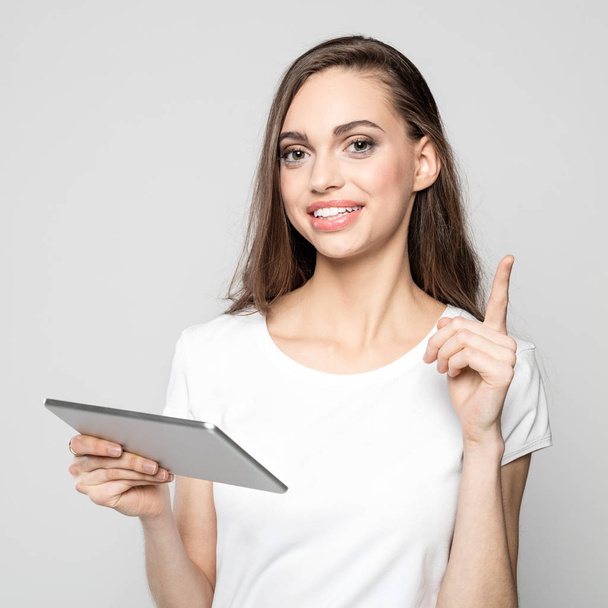 Portrait of beautiful long hair female student wearing white t-shirt, holding a digital tablet and pointing with index finger. Studio shot against grey background. - Photo, Image