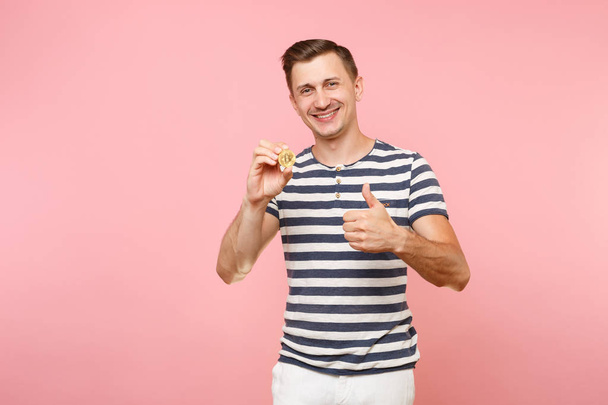 Portrait of smiling young man in striped t-shirt holding bitcoin, metal coin of golden color isolated on pink background. People sincere emotions lifestyle concept. Advertising area. Future currency - Valokuva, kuva