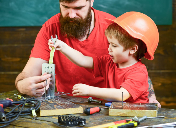 Father with beard teaching little son to use tools in classroom, chalkboard on background. Boy, child busy in protective helmet makes by hand, repairing, work with tools with dad. Team work concept - Фото, изображение