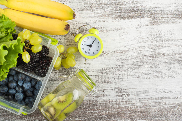 Alarm clock and berries in lunch box and fruits on wooden table - Photo, Image