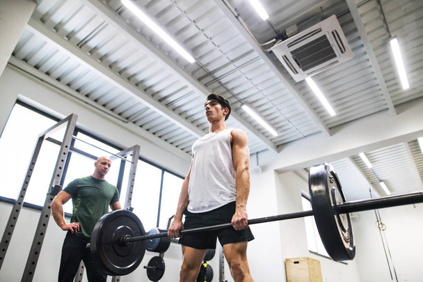 Fit young man with a personal trainer in gym working out, lifting barbell. - Photo, image