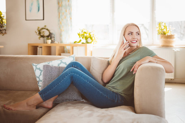 Indoor shot of happy, beautiful young woman relaxing on sofa in the modern living room and talking on smart phone. - Foto, Imagem