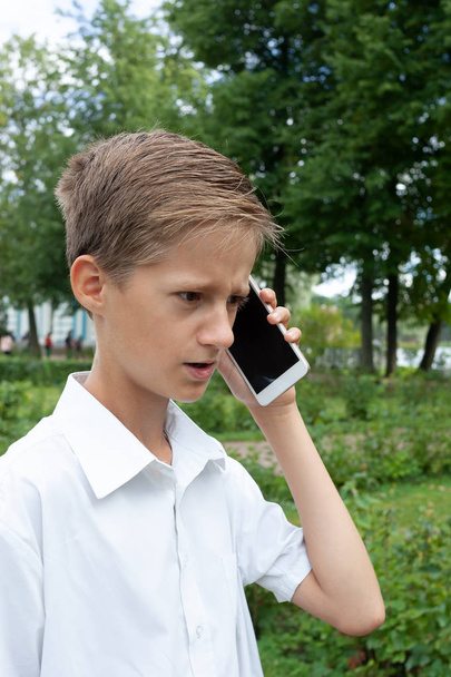 the boy emotionally speaks in the park by phone in summer day - Фото, изображение