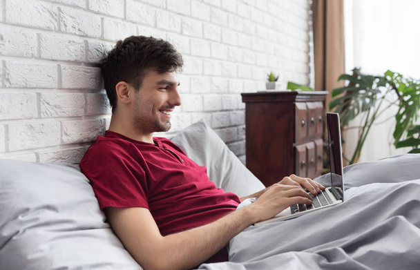 Joyeux bel homme en utilisant un ordinateur portable au lit
 - Photo, image
