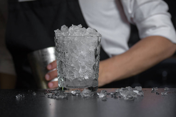 Imagem de close-up de um copo cheio de gelo acidente, de pé no balcão da barra, bartender detém shaker refrigerado no fundo
.  - Foto, Imagem