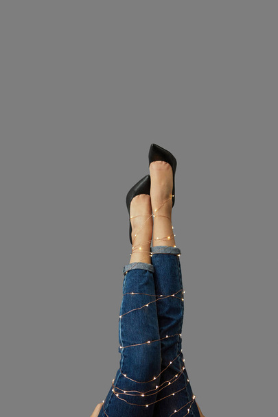 A girls legs tied with a Christmas garland in shoes and jeans on a gray background with copy space. - Fotó, kép