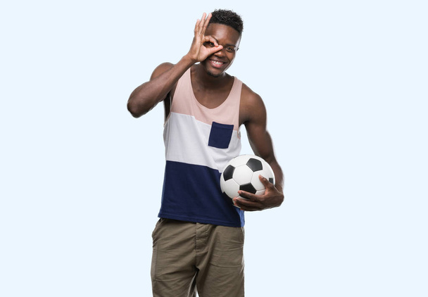 Jonge african american man die voetbal voetbal met blij gezicht lachend doen ok teken met de hand op oog kijken door de vingers - Foto, afbeelding