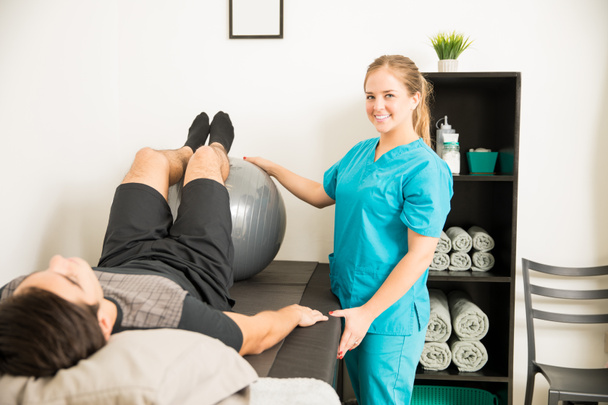 Χαμογελαστός νεαρός φυσική θεραπευτής χρησιμοποιώντας μπάλα γυμναστικής για προπόνηση τα πόδια του ασθενούς στην κλινική - Φωτογραφία, εικόνα