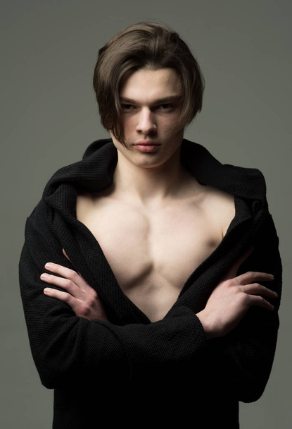 Man with smooth skin on face, neck and chest looking at camera. Guy on confident face in black bathrobe holds arms folded, grey background. Macho with stylish long haircut. Masculinity concept. - Foto, afbeelding