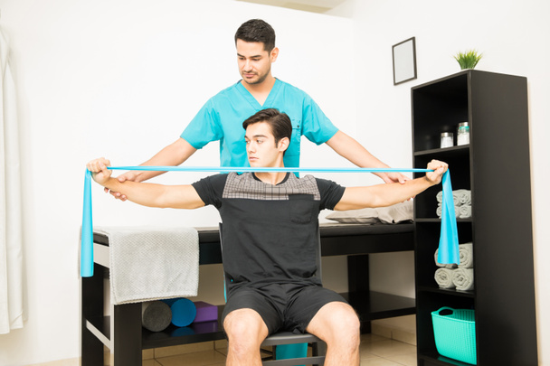 Physiotherapeut hilft männlichen Patienten mit Resistenzband im Krankenhaus - Foto, Bild