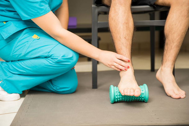 Χαμηλό τμήμα γυναικείας θεραπευτή βοηθώντας ασθενή για να χρησιμοποιήσει τα πόδια ρολό για πελματικό fasciitis και τον πόνο ανακούφιση στο νοσοκομείο - Φωτογραφία, εικόνα