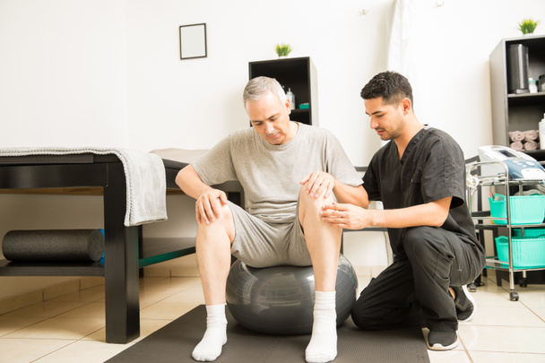 Fizikai terapeuta segíti, érett férfi beteg ül a gyakorlat labda klinikán - Fotó, kép