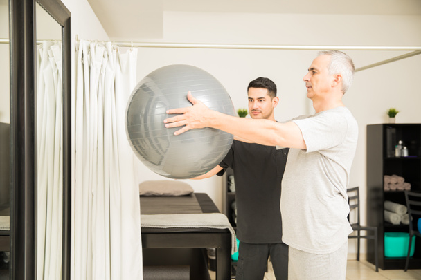 Focused physiotherapist assisting elderly man lifting exercise ball in hospital - Фото, изображение