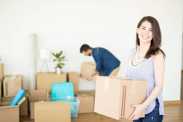 glückliche Frau trägt Schachtel, während Mann im Hintergrund Arrangement im neuen Zuhause macht - Foto, Bild