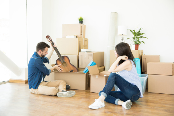 mies poistaa kitaran liikkuvasta laatikosta istuessaan kovapuulattialla naisen kanssa uudessa kodissa
 - Valokuva, kuva