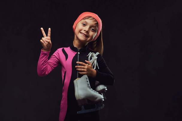 Happy little skater girl dressed in sportswear holds ice skates on a shoulder and shows victory hand. Isolated on dark textured background. - Photo, Image