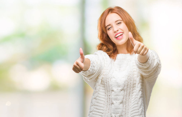 Young beautiful woman over isolated background wearing winter sweater approving doing positive gesture with hand, thumbs up smiling and happy for success. Looking at the camera, winner gesture. - Photo, Image