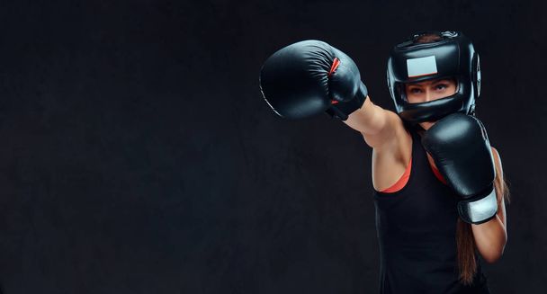 Mujer deportiva en ropa deportiva con un casco protector y guantes de boxeo, entrenamiento en el gimnasio. Aislado sobre un fondo de textura oscura
. - Foto, Imagen