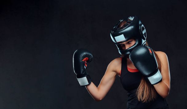 Mulher esportiva em roupas esportivas usando um capacete protetor e luvas de boxe, treinando no ginásio. Isolado em um fundo texturizado escuro
. - Foto, Imagem