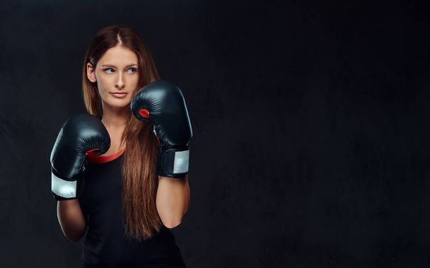 Mujer deportiva vestida con ropa deportiva con guantes de boxeo posando en un estudio. Aislado sobre un fondo de textura oscura
. - Foto, Imagen