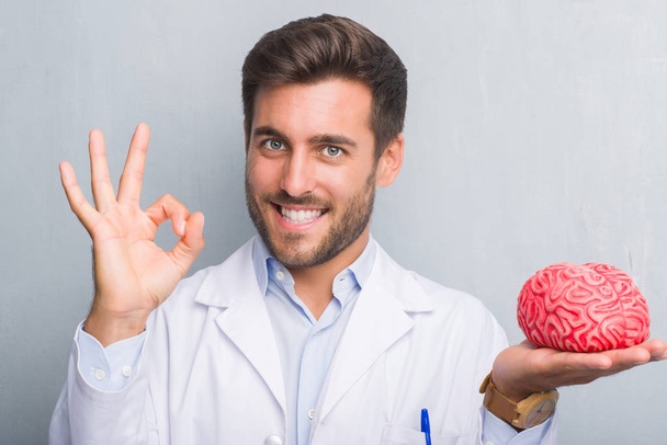 Handsome young doctor psychologist man over grey grunge wall holding a brain doing ok sign with fingers, excellent symbol - Foto, imagen