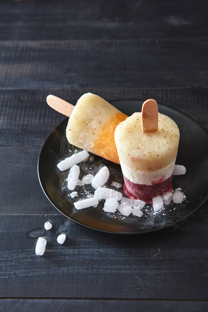 Homemade smoothies lolly on a stick. Multicolored berry, coffee ice cream with ice cubes in a black plate on a dark wooden background with space for text. Flat lay - Фото, изображение