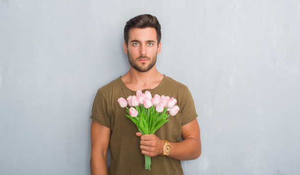 Красивий молодий чоловік над сірою гранжевою стіною тримає букет квітів з впевненим виразом на розумному обличчі думаючи серйозно
 - Фото, зображення