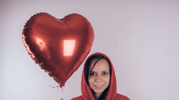 A girl in a red sweatshirt in her hand a red balloon in the form of a heart. Student posing on white background - Fotografie, Obrázek