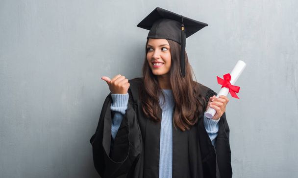 Young brunette woman over grunge grey wall wearing graduate uniform holding degree pointing and showing with thumb up to the side with happy face smiling - Foto, Imagen