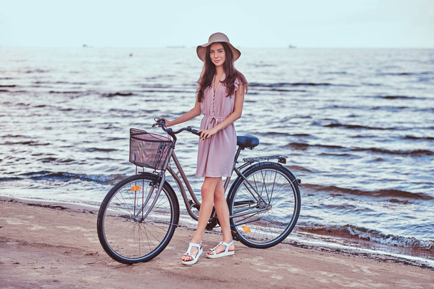 Happy sensual girl in hat wearing dress walks with her bicycle on the beach against amazing seaside background. - Photo, Image