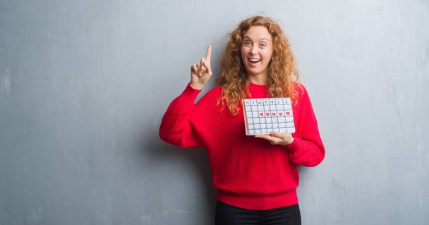 Young redhead woman over grey grunge wall holding period calendar surprised with an idea or question pointing finger with happy face, number one - Foto, Bild