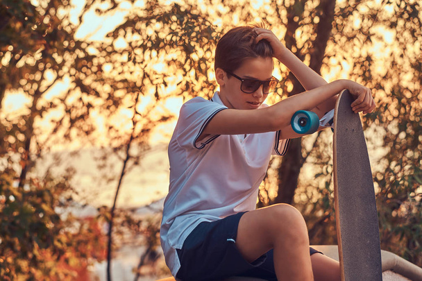 Jonge skater jongen in zonnebril gekleed in t-shirt en shorts zitten op de stenen vangrail buitenshuis bij een zonsondergang.  - Foto, afbeelding
