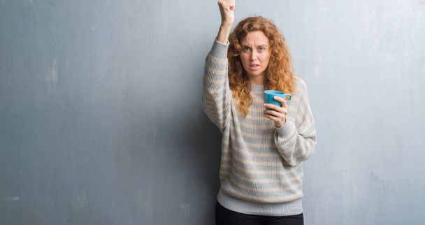 Jonge roodharige vrouw over grijs grunge wall drinken een kopje koffie boos en gefrustreerd schreeuwen met woede, gek en schreeuwen met opgeheven hand, woede concept - Foto, afbeelding