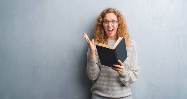 Jeune femme rousse sur un mur gris grunge lisant un livre très heureux et excité, expression gagnante célébrant la victoire en criant avec un grand sourire et les mains levées
 - Photo, image