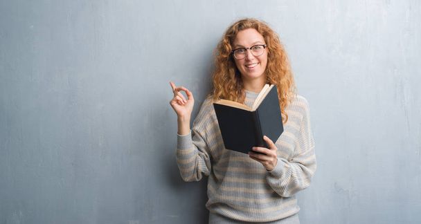 Jeune femme rousse sur un mur gris grunge lisant un livre très heureux pointant avec la main et le doigt sur le côté
 - Photo, image