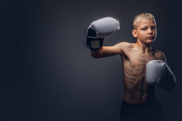 Boxeador joven sin camisa con pelo rubio usando guantes de boxeo muestra un gancho de boxeo
. - Foto, imagen