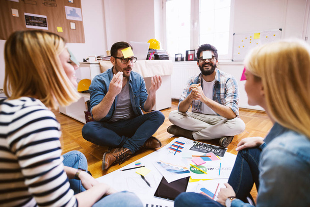 сотрудники наслаждаются свободным временем и поднимают командный дух, сидя в кругу на полу и играя в игры с бумажными наклейками на лбу
 - Фото, изображение