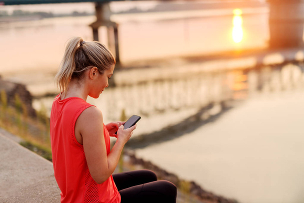 Blond beautiful girl in sports clothes sitting near river and checking her telephone. - Photo, Image
