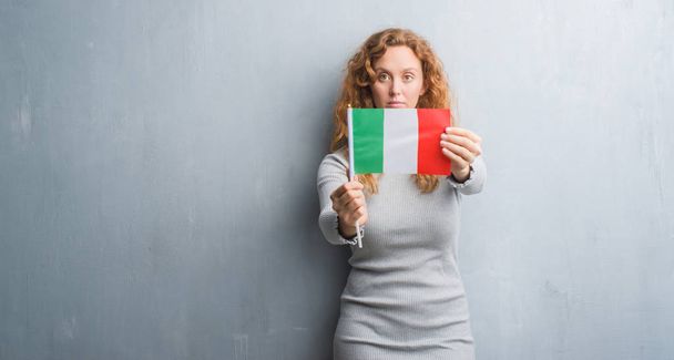 Giovane rossa donna oltre grigio grunge muro tenendo bandiera d'Italia con un'espressione fiduciosa sul viso intelligente pensando grave
 - Foto, immagini