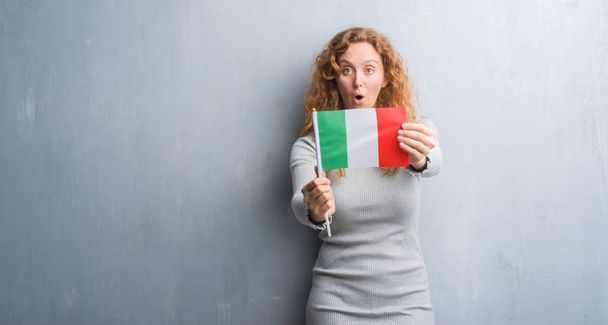 Молодая рыжая женщина над серой гранж-стеной с флагом Италии, напуганная удивленным лицом, напуганная и взволнованная выражением страха
 - Фото, изображение