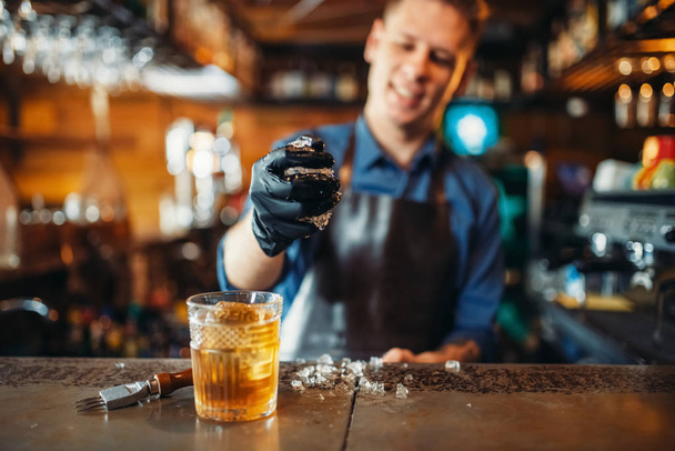 Μπάρμαν αρσενικό στην ποδιά λειτουργεί με πάγο στο μπαρ του μετρητή. Αλκοόλ Παρασκευή αφεψήματος. Επάγγελμα μπάρμαν - Φωτογραφία, εικόνα