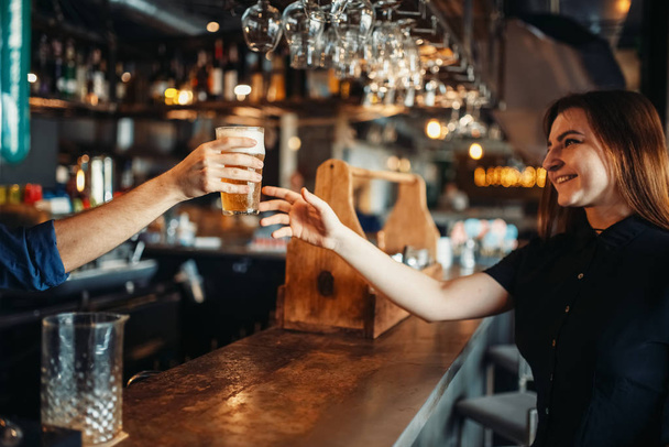 Μπάρμαν αρσενικό περνά ένα ποτήρι μπίρα θηλυκό επισκέπτη. Επάγγελμα μπάρμαν μπάρμαν και γυναίκα στο μπαρ του μετρητή - Φωτογραφία, εικόνα