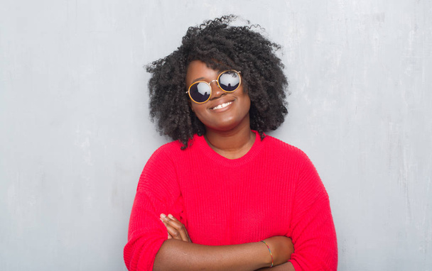 Νεαρός αφρικανική αμερικανική συν μέγεθος γυναίκα πάνω από τοίχο γκρι grunge, φορώντας retro γυαλιά ηλίου χαρούμενο πρόσωπο χαμογελά με σταυρωμένα χέρια, κοιτάζοντας την κάμερα. Θετικό πρόσωπο. - Φωτογραφία, εικόνα