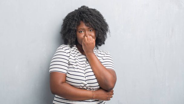 Jeune femme afro-américaine plus la taille sur le mur gris grunge regardant stressé et nerveux avec les mains sur les ongles mordants de la bouche. Problème d'anxiété
. - Photo, image